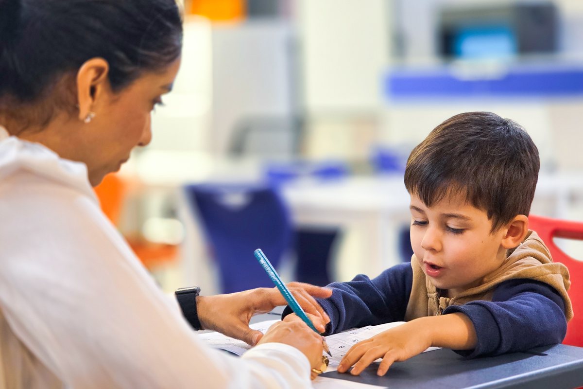 Mother or teacher helping two young girls with art or homework at a white desk.
